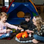 Three 4K students sitting around a play fire pretending to roast marshmallows.